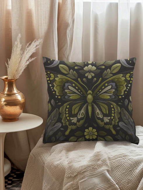 A square decorative pillow featuring an intricately detailed butterfly design in olive green, sage, and cream colors against a black background. The butterfly is surrounded by botanical elements with symmetrical floral patterns.